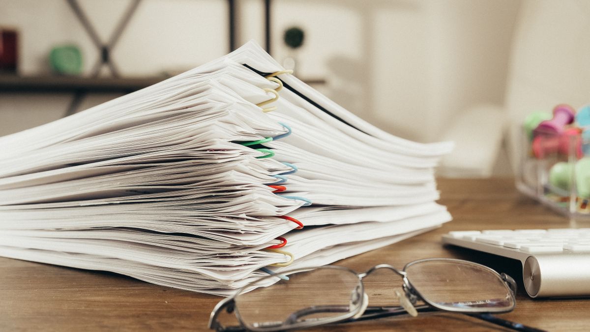 A desk covered in stacked paper forms and documents, the kind of manual paperwork that slows credentialing down